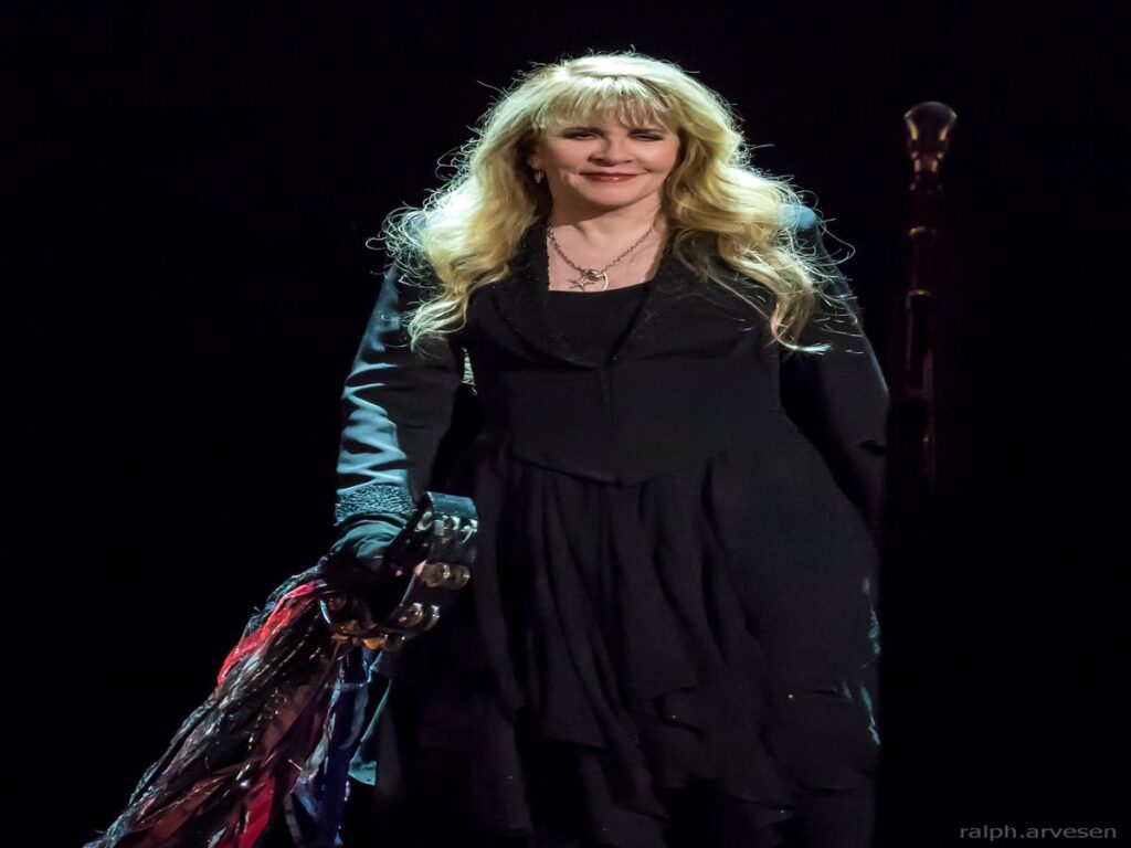 Stevie Nicks performing on stage, wearing a black outfit and holding a tambourine, smiling at the audience.