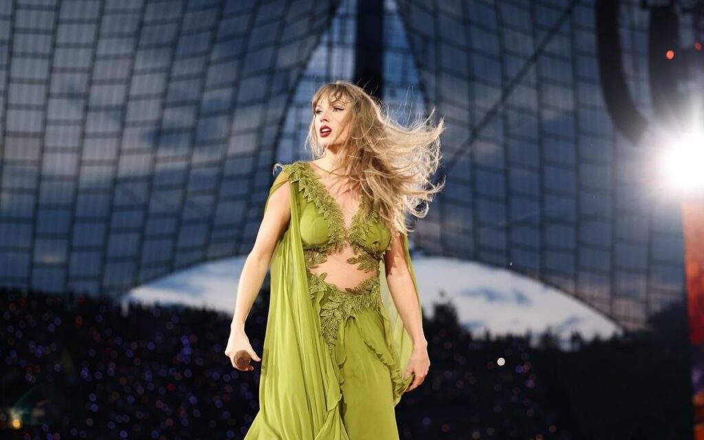 "Taylor Swift performs on stage, wearing a stunning green gown with intricate details, her hair flowing as she captivates the audience under a beautifully lit backdrop."