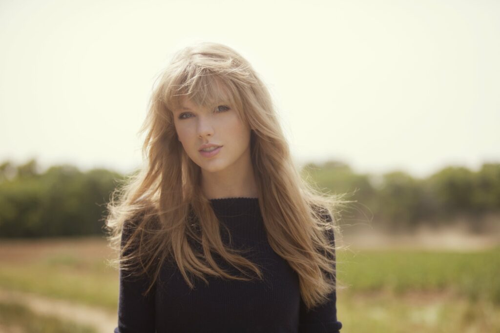 A young woman with long, flowing hair stands in a sunlit field, wearing a dark sweater. She gazes softly at the camera.