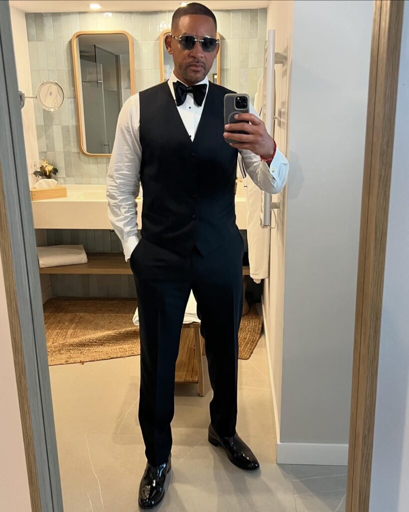 Will Smith stands in front of a mirror, wearing a black tuxedo with a bow tie and sunglasses. He poses confidently, showcasing his stylish look in a well-lit bathroom setting.