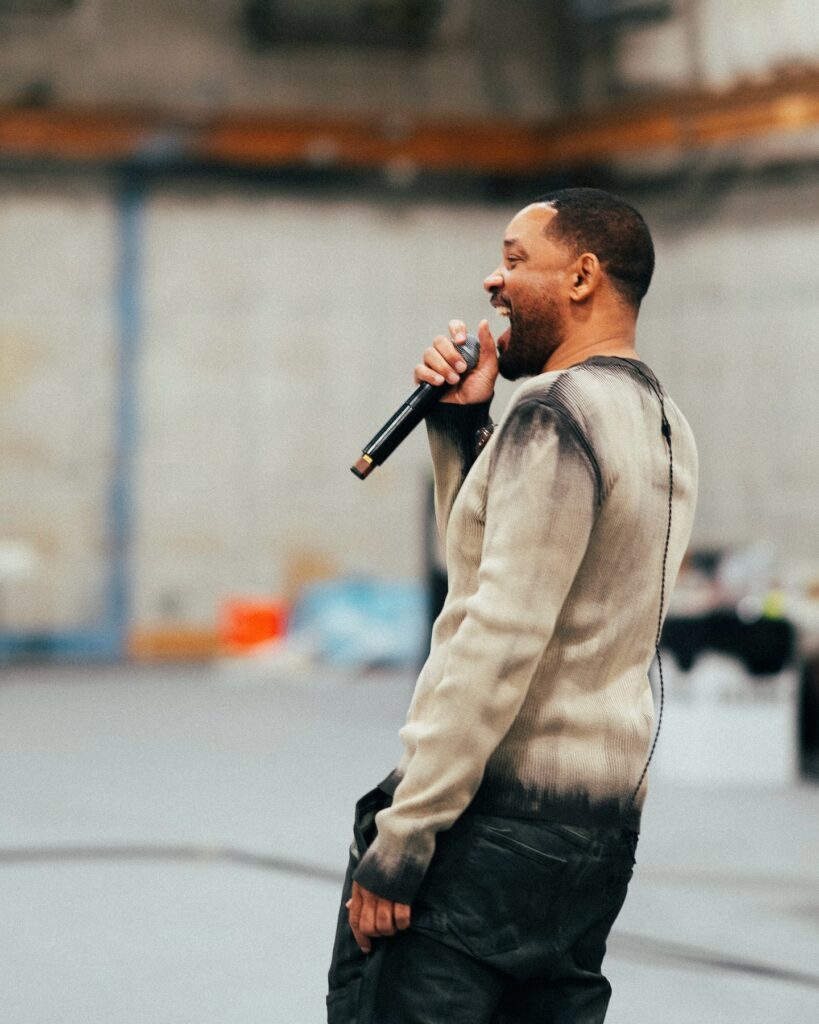 Will Smith laughing and speaking into a microphone while wearing a stylish gray sweater and black pants.