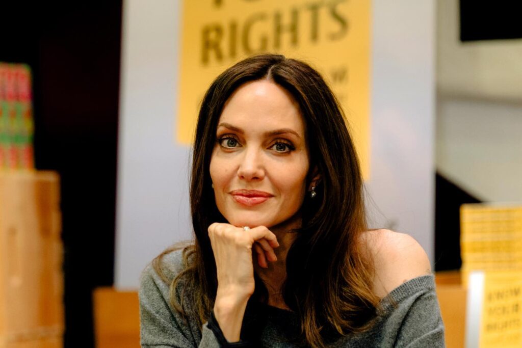 Angelina Jolie poses with a book at an event, showcasing her thoughtful expression, wearing a gray sweater.