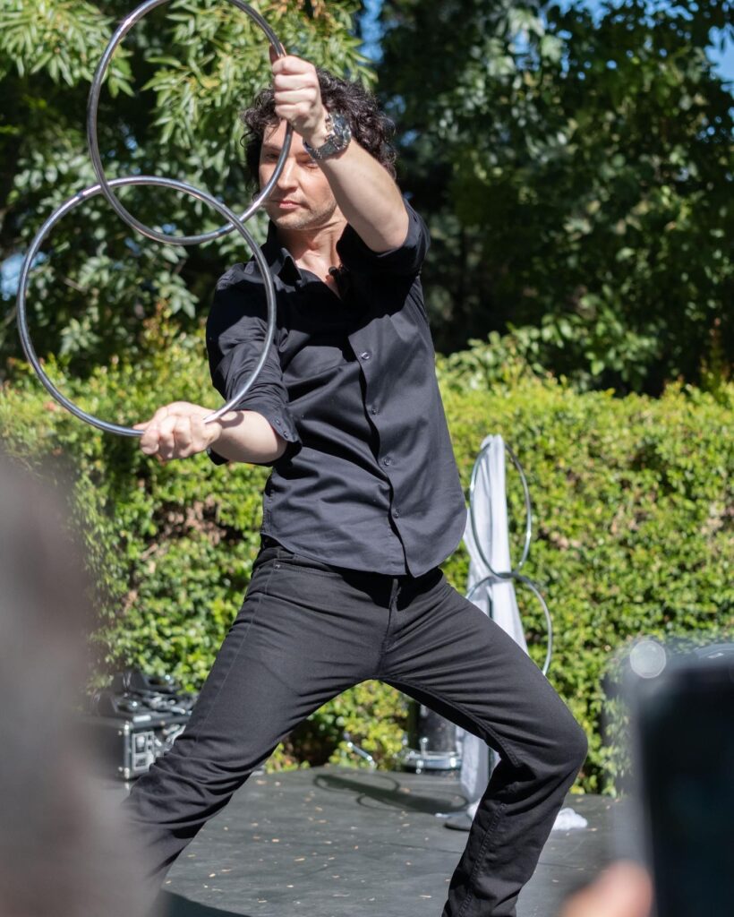 A performer skillfully manipulates two shiny metal hoops in a dynamic pose, dressed in a black shirt and pants, outdoors.