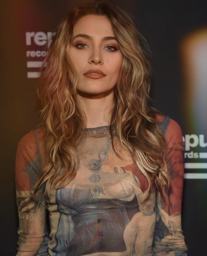 Paris Jackson poses confidently with tousled hair, wearing a unique shirt featuring artistic designs, against a dark backdrop.