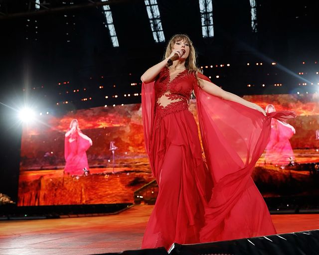 Taylor Swift performs on stage in a stunning red dress, with flowing fabric and a microphone, in front of a vibrant backdrop.