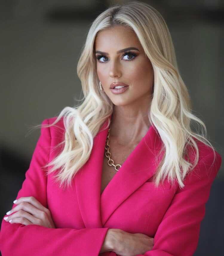 Emma Hernan poses confidently in a vibrant pink blazer with her arms crossed, showcasing her styled blonde hair and glamorous makeup.