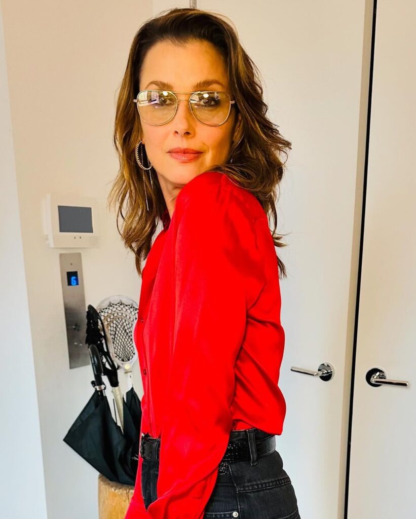 Bridget Moynahan in a red blouse and glasses, looking over her shoulder.