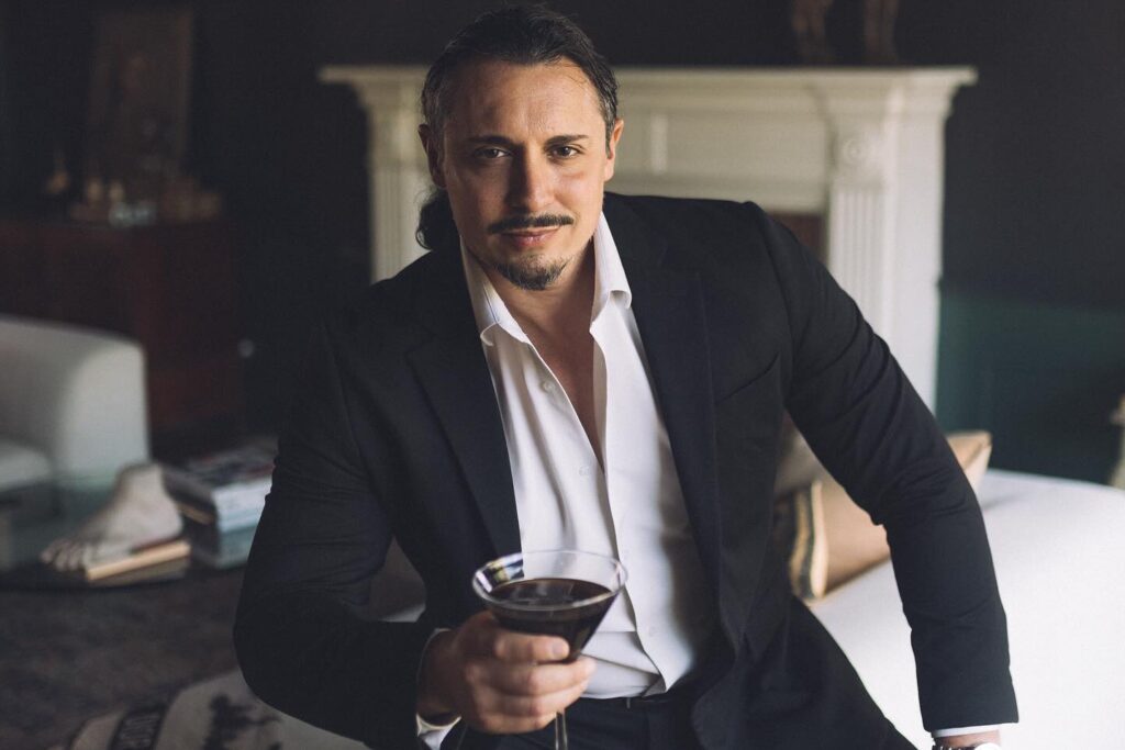 Peter Madrigal seated on a couch, wearing a black suit with an open collar, holding a cocktail glass.