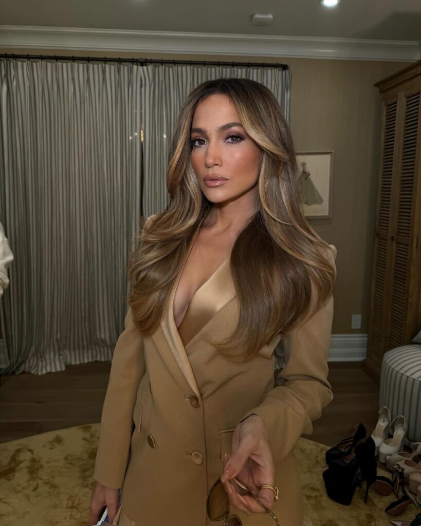 : In this image, Jennifer Lopez presents a glamorous appearance with long, flowing hair and a fashionable beige blazer. The background features soft curtains and a collection of shoes, enhancing the stylish ambiance.