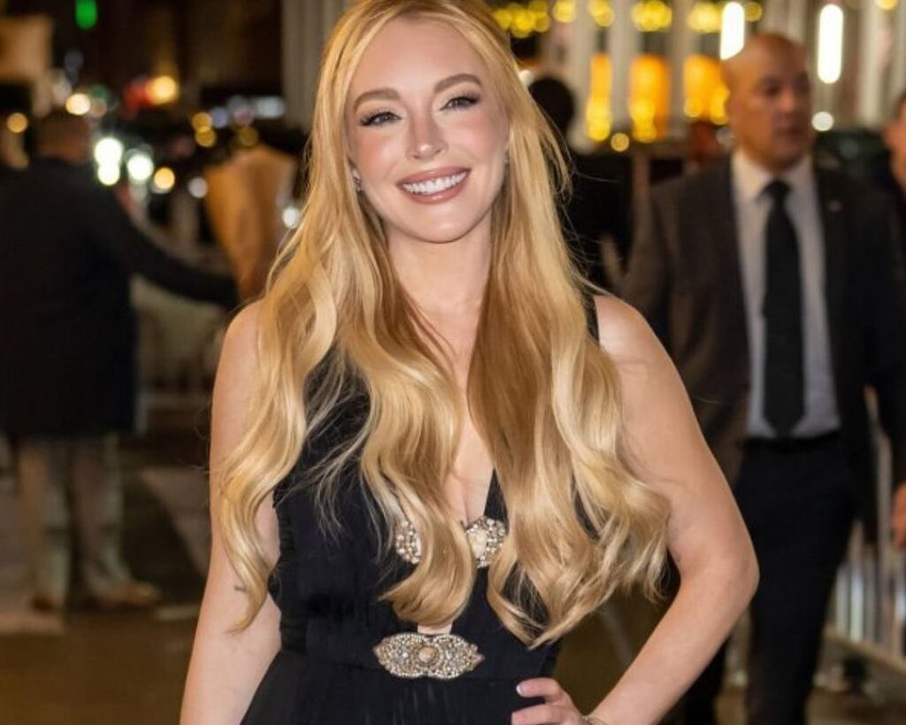 Lindsay Lohan smiling in a black dress with long hair, posing confidently at an event.