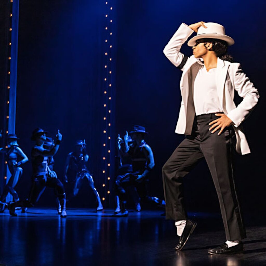 A performer dressed as Michael Jackson showcases a dance move in a dramatic spotlight, surrounded by backup dancers in dark outfits.