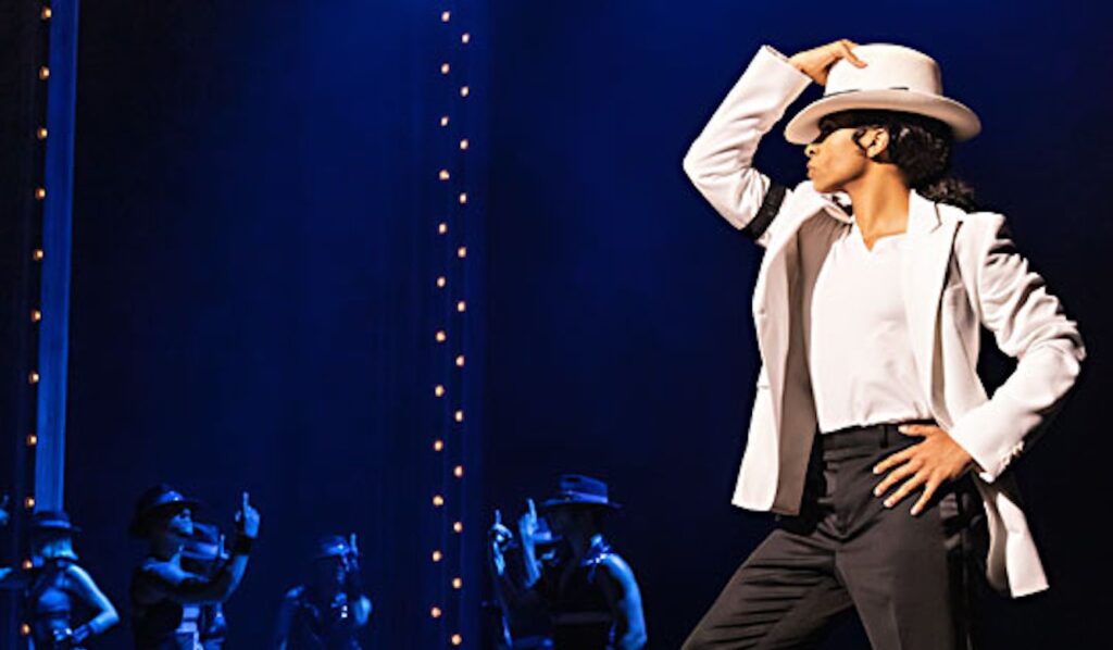 A performer dressed as Michael Jackson showcases a dance move in a dramatic spotlight, surrounded by backup dancers in dark outfits.