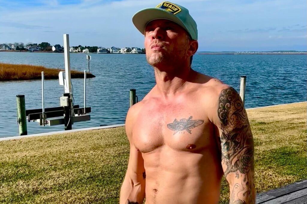 Ryan Phillippe stands shirtless by the water, showing off his tattoos and wearing teal joggers and a cap, with a serene landscape behind him.