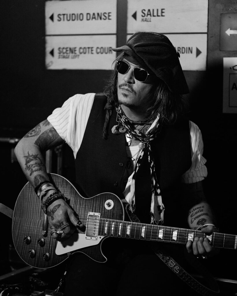 Musician Johnny depp wearing hat and sunglasses plays an electric guitar backstage near French direction signs, black‑and‑white photo.