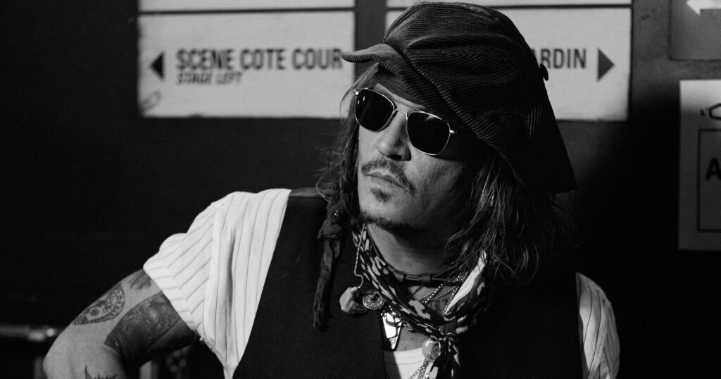 Johnny depp wearing hat and sunglasses plays an electric guitar backstage near French direction signs, black‑and‑white photo.