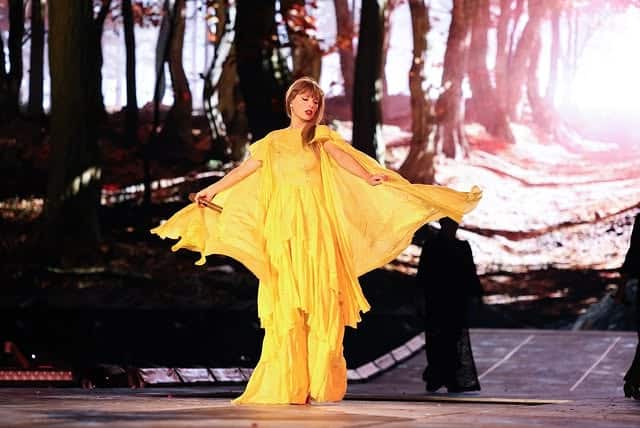 Performer in a flowing yellow gown stands onstage in a forest‑like set, arms extended as light filters through trees.