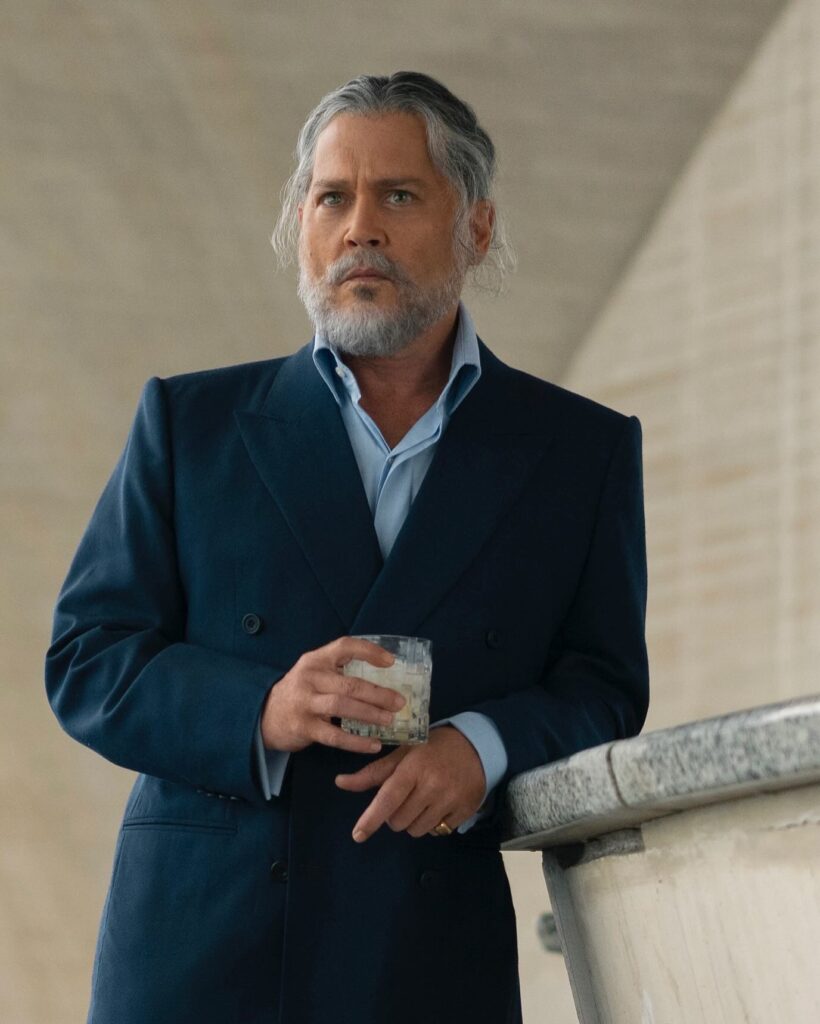Johnny depp with a trimmed beard in a dark double‑breasted suit holds a glass with ice while standing by a curved stone railing in a modern, minimalist interior.