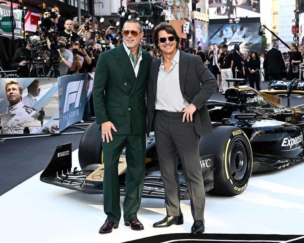 Two men in suits pose by a black‑and‑gold Formula 1 car at a premiere.