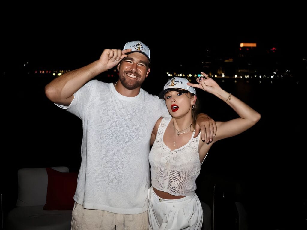 Taylor and Travis in white outfits and matching caps pose arm‑in‑arm at night on a waterfront with city lights in the background.