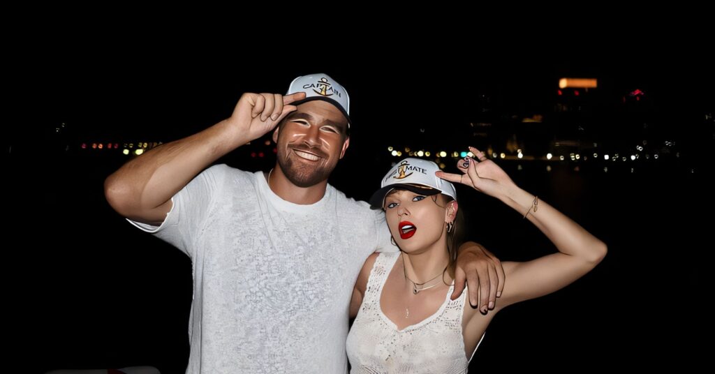 A smiling couple in white outfits and matching caps pose arm‑in‑arm at night on a waterfront with city lights in the background.