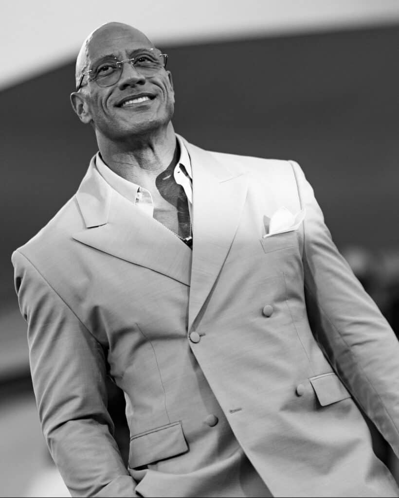 Black‑and‑white portrait of a smiling bald person in a double‑breasted suit with pocket square, looking upward at an event.