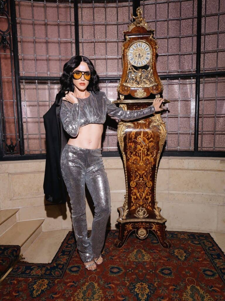 Katy Perry Woman in shimmering silver crop top and pants, yellow sunglasses, standing beside an ornate grandfather clock in a vintage room.