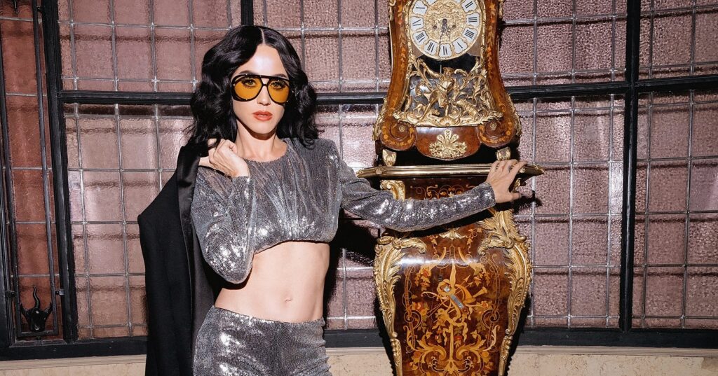 Katy Perry in shimmering silver crop top and pants, yellow sunglasses, standing beside an ornate grandfather clock in a vintage room.