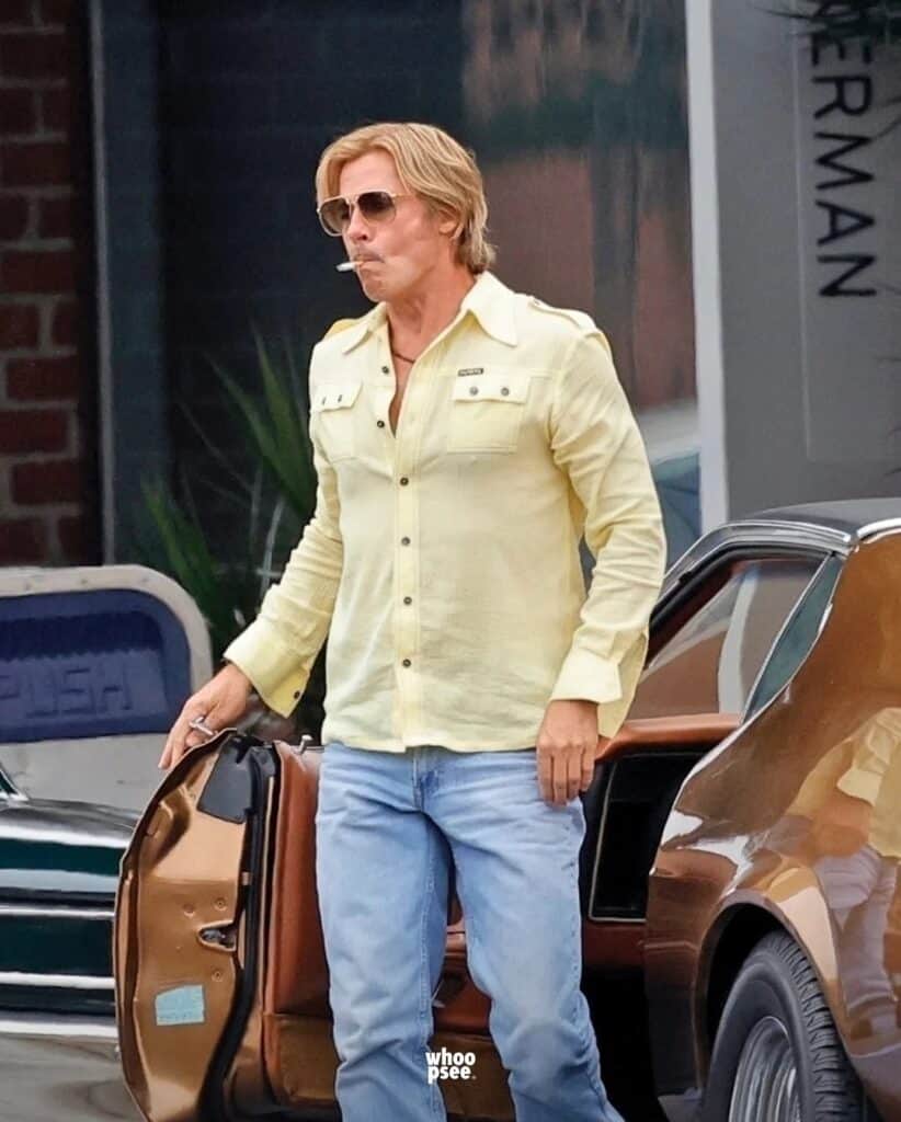 Bradpit in yellow shirt and jeans exits vintage car, cigarette in mouth, wearing aviator sunglasses on city street.