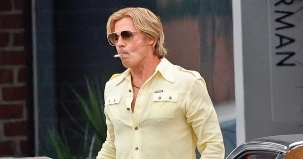 Man in yellow shirt and jeans exits vintage car, cigarette in mouth, wearing aviator sunglasses on city street.