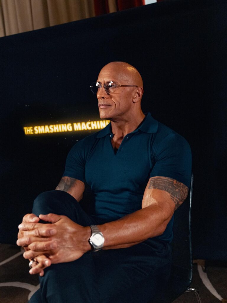 Dwyane johnson Bald muscular person wearing glasses and a dark polo shirt sits cross‑legged, hands clasped, against a black backdrop reading “The Smashing Machine,” warm indoor lighting.