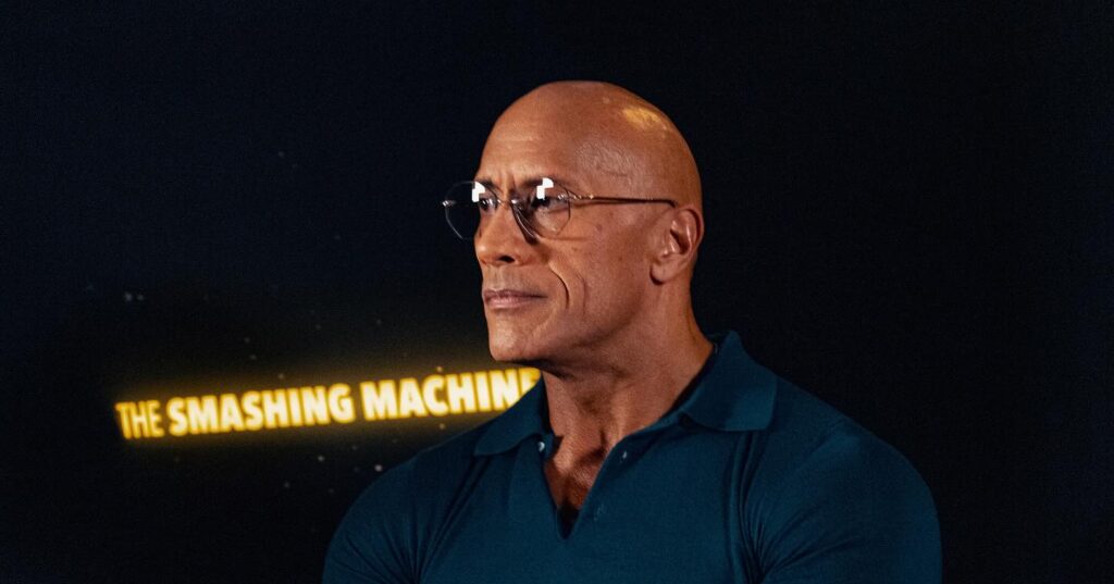 Dwyane johnson Bald muscular person wearing glasses and a dark polo shirt sits cross‑legged, hands clasped, against a black backdrop reading “The Smashing Machine,” warm indoor lighting.
