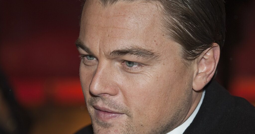 Leonardo DiCaprio Close-up of a man in formal attire at an indoor event, warm red-orange blurred background.