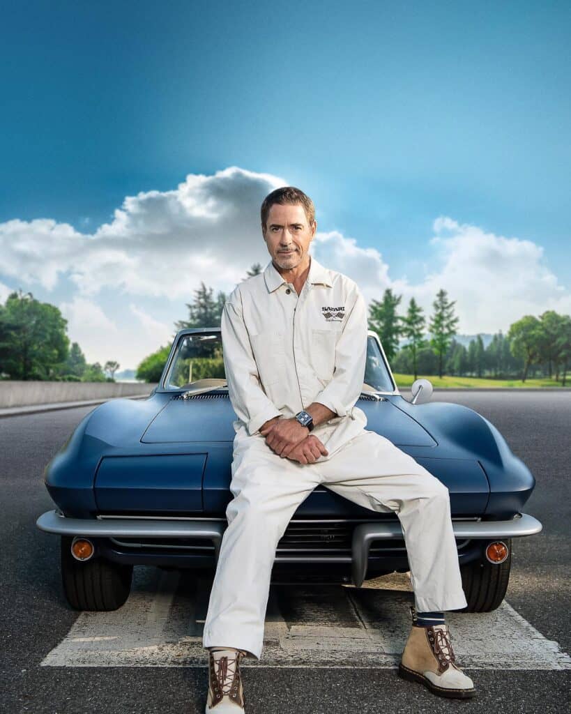 Robert Downey Jr., dressed in a white jumpsuit, sits on the hood of a classic blue convertible. He is on a road with a starting line, with a background of green trees and a partly cloudy blue sky.