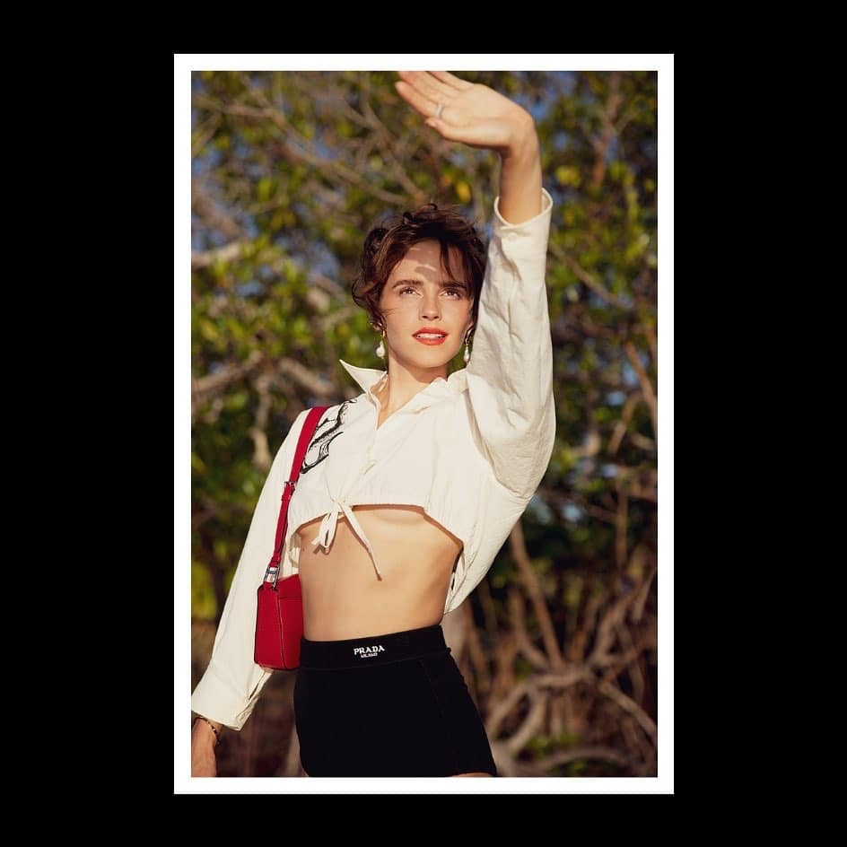 Emma Watson stands outdoors in front of leafy trees, shielding her eyes from the sun with one hand raised. She is wearing a white long-sleeved crop top, black high-waisted shorts with a Prada logo, and a red shoulder bag.