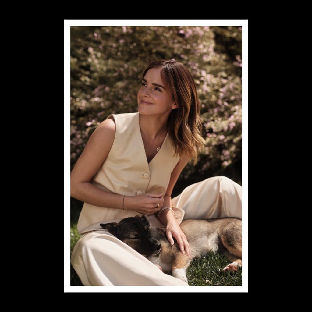 Emma Watson is sitting on the grass, smiling and looking away from the camera. She is wearing a cream-colored vest and pants, and is gently petting a sleeping puppy in her lap. The background is a soft-focus shot of pink flowers.