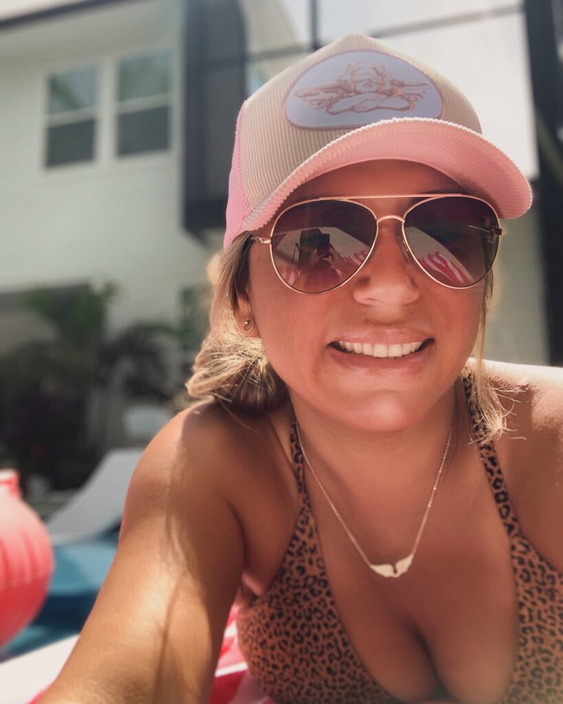 Jennifer Lawrence person in a pink cap and sunglasses lounging poolside in a patterned swimsuit, taking a sunny selfie.
