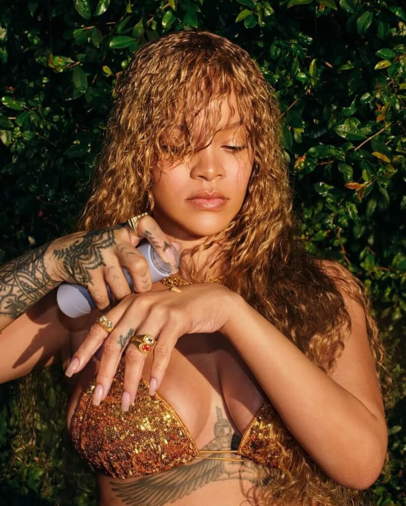 A medium shot of Rihanna outdoors, standing in front of lush green foliage. Her eyes are closed as she sprays a product from a light purple bottle onto her décolletage. She has long, curly, honey-blonde hair with bangs falling across her face. She is wearing a gold sequined bikini top, several gold rings with gemstones, and gold necklaces. Her extensive tattoos are visible on her hands, forearms, and ribcage. The lighting is bright and warm, highlighting her glowing skin.