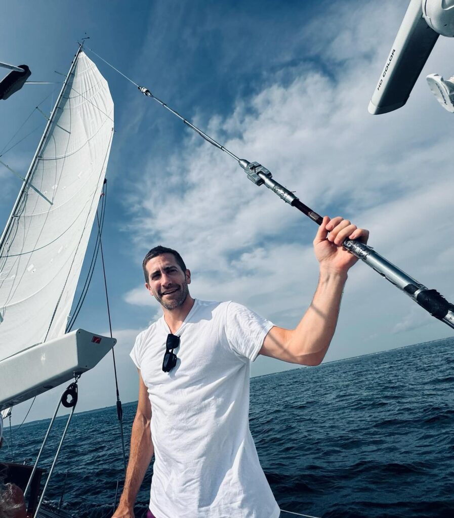 Jake Gyllenhaal in a white T‑shirt stands on a sailboat, holding a rigging line against a blue, partly cloudy sky. Sunglasses hang from his collar as waves stretch to the horizon, capturing a relaxed, adventurous moment on open water.