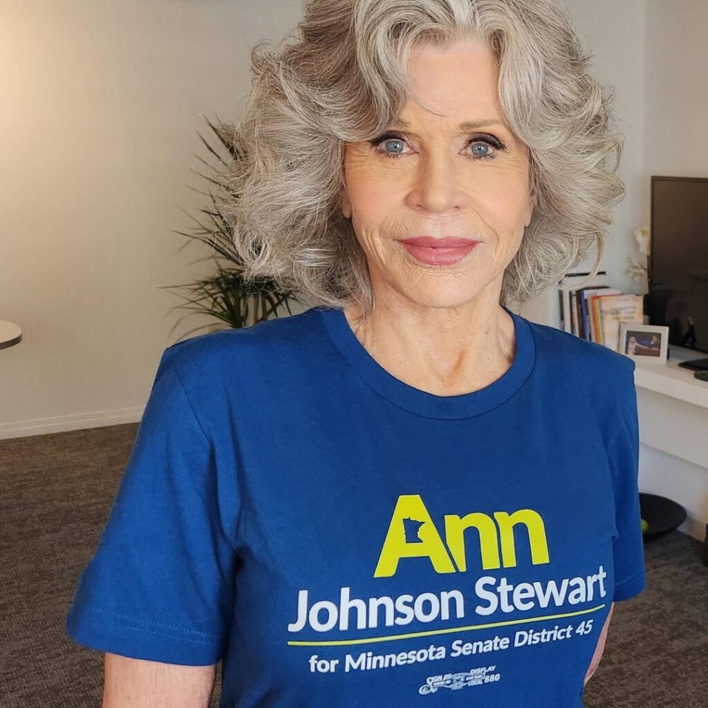 Jane Fonda haired adult in a blue campaign T‑shirt stands indoors, looking at the camera with a calm expression.