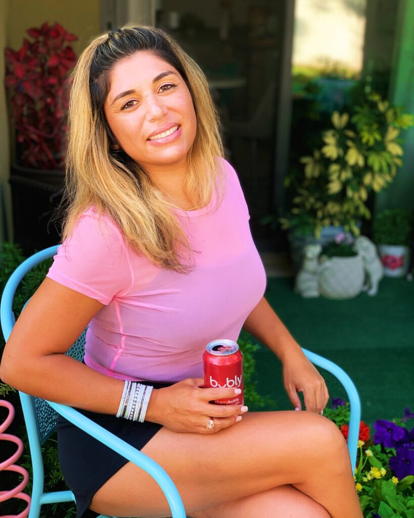 Jennifer Lawrence seated outdoors on a blue chair holding a red sparkling water can, wearing a pink top and smiling.