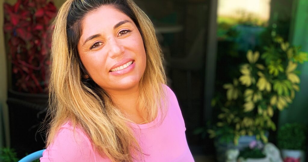Jennifer Lawrence seated outdoors on a blue chair holding a red sparkling water can, wearing a pink top and smiling.
