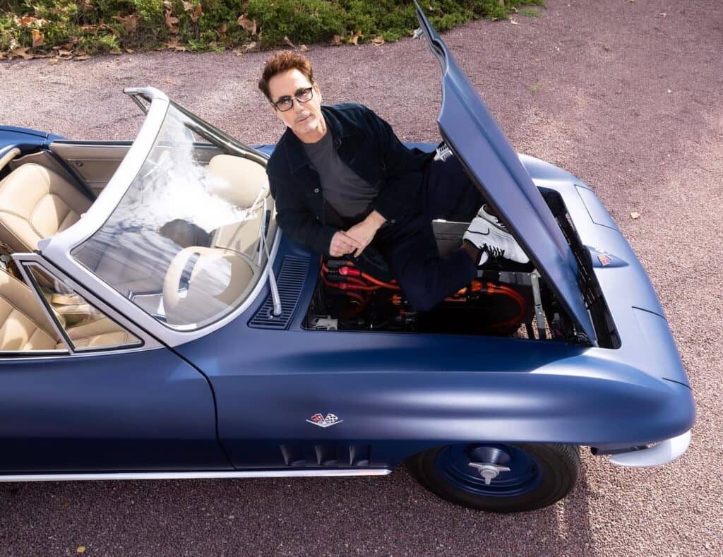 A high-angle shot of Robert Downey Jr. sitting inside the engine bay of a classic matte blue convertible car with its hood open. He is wearing glasses and a dark jacket, looking up at the camera.