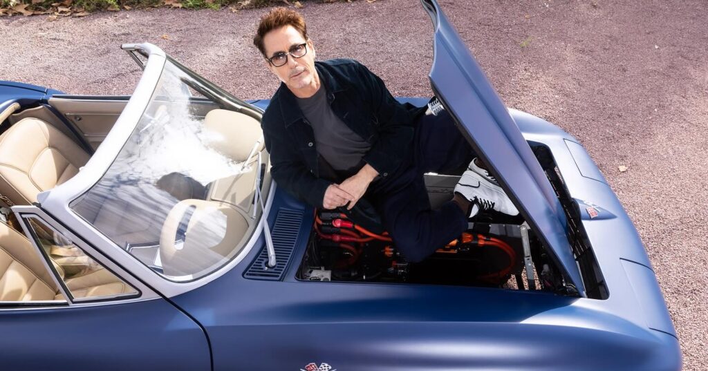 A high-angle shot of Robert Downey Jr. sitting inside the engine bay of a classic matte blue convertible car with its hood open. He is wearing glasses and a dark jacket, looking up at the camera.