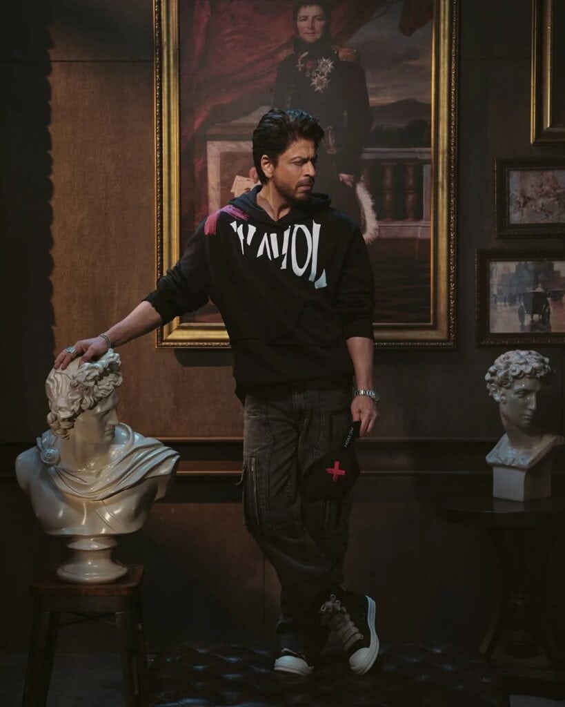 Shah Rukh Khan in a black graphic hoodie and cargo pants, standing in a dimly lit room with classical paintings and marble busts.