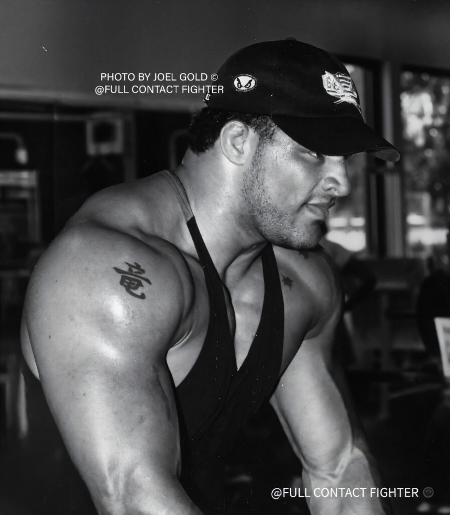 Mark Kerr in a gym wearing a tank top and cap, showing shoulder tattoo, photographed in black and white during an intense workout.