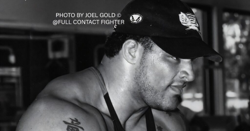Mark Kerr in a gym wearing a tank top and cap, showing shoulder tattoo, photographed in black and white during an intense workout.