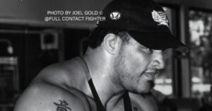 Mark Kerr in a gym wearing a tank top and cap, showing shoulder tattoo, photographed in black and white during an intense workout.
