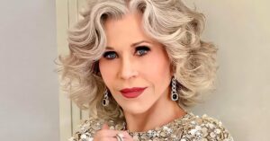 Elegant older adult with silver curls wearing a beaded floral top, posing indoors with jewelry and soft makeup.