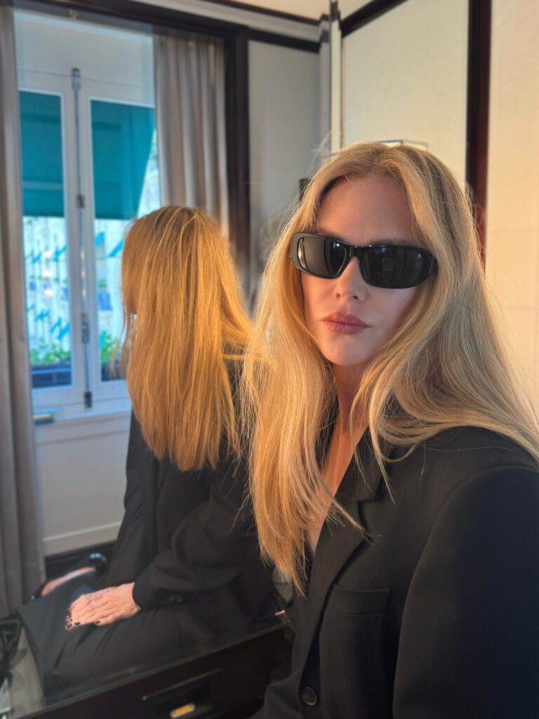 Nicole Kidman in black blazer and dark sunglasses sits by a mirror, long blonde hair visible front and reflection, soft indoor light and window in background.