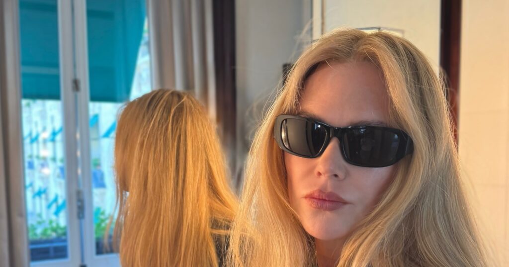 Nicole Kidman in black blazer and dark sunglasses sits by a mirror, long blonde hair visible front and reflection, soft indoor light and window in background.